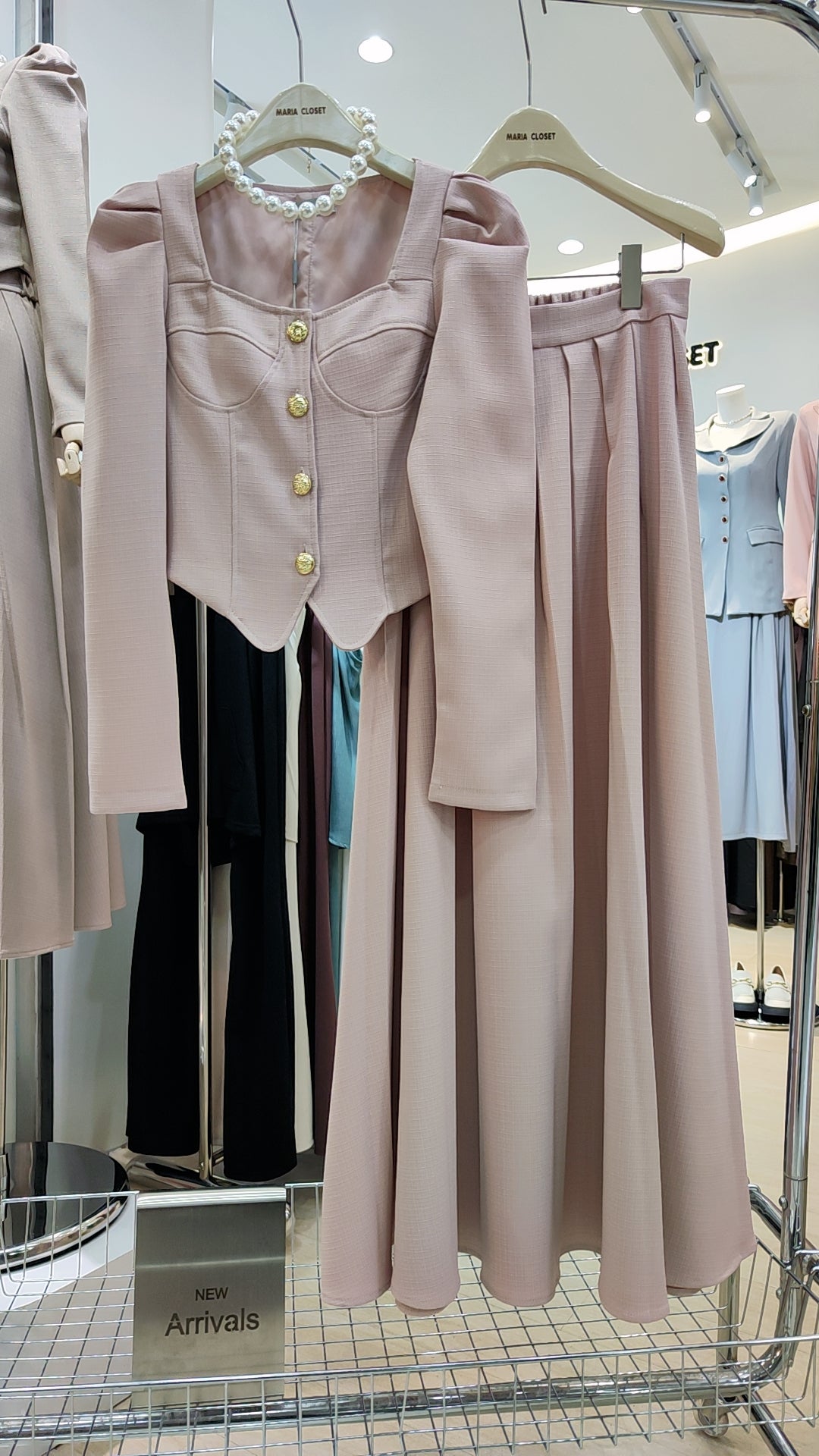The "Flowy Tweed" Co-ord Set in soft pink with a fitted top and flowing long skirt on hangers in a boutique.